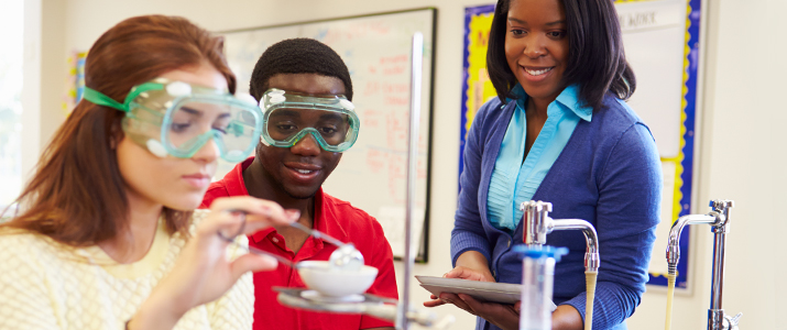 Two students in a classroom lab with safety goggles on performing a science experiment overseen by a teacher figure.